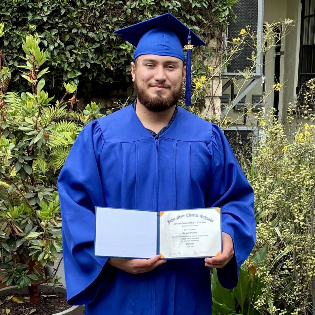 German is wearing a blue graduation cap and gown and holds up his diploma in front of greenery