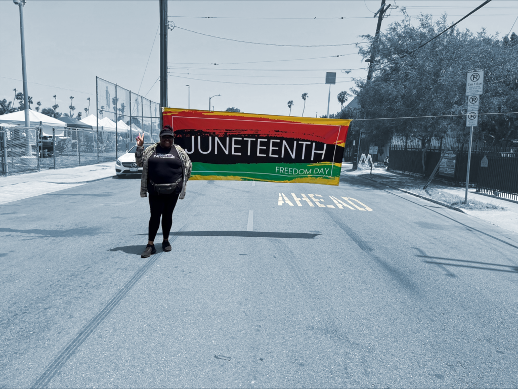 Tanisha in front of Juneteenth banner across street next to park. Festival tents are lined up in the park.